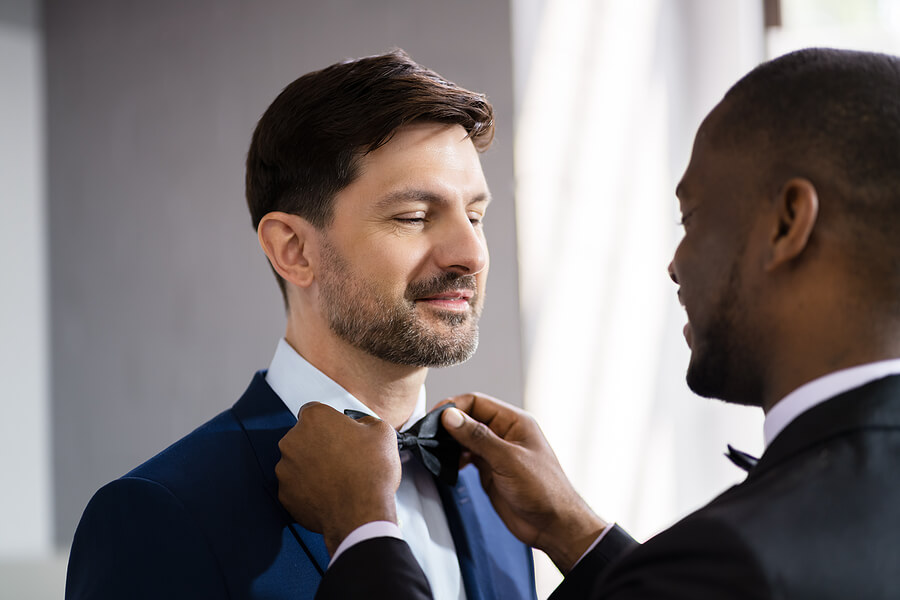 Two grooms at their wedding, with the Black groom straightening the white groom's bowtie